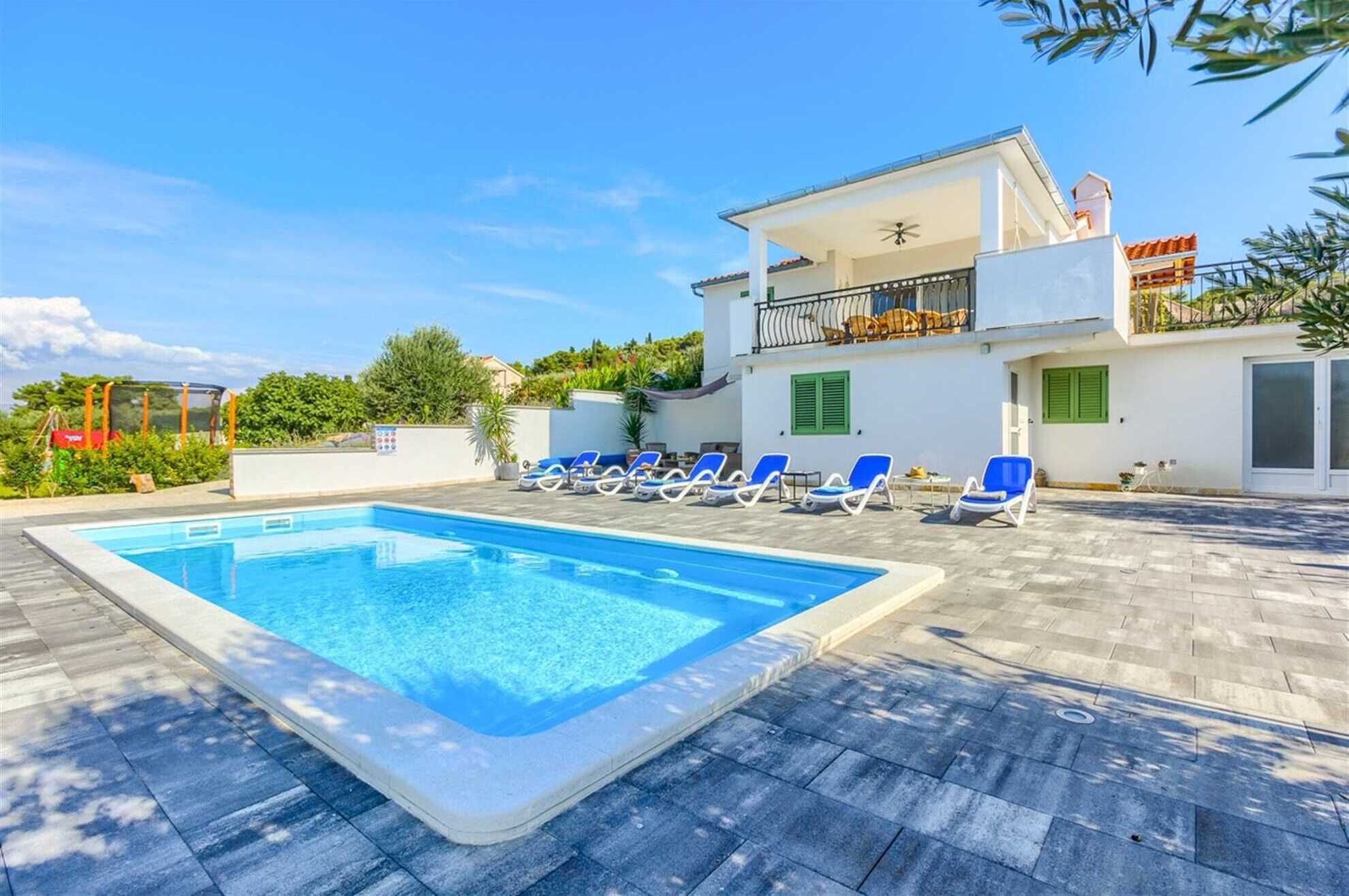 A swimming pool with lounge chairs, a white building with a balcony, and greenery in the background.