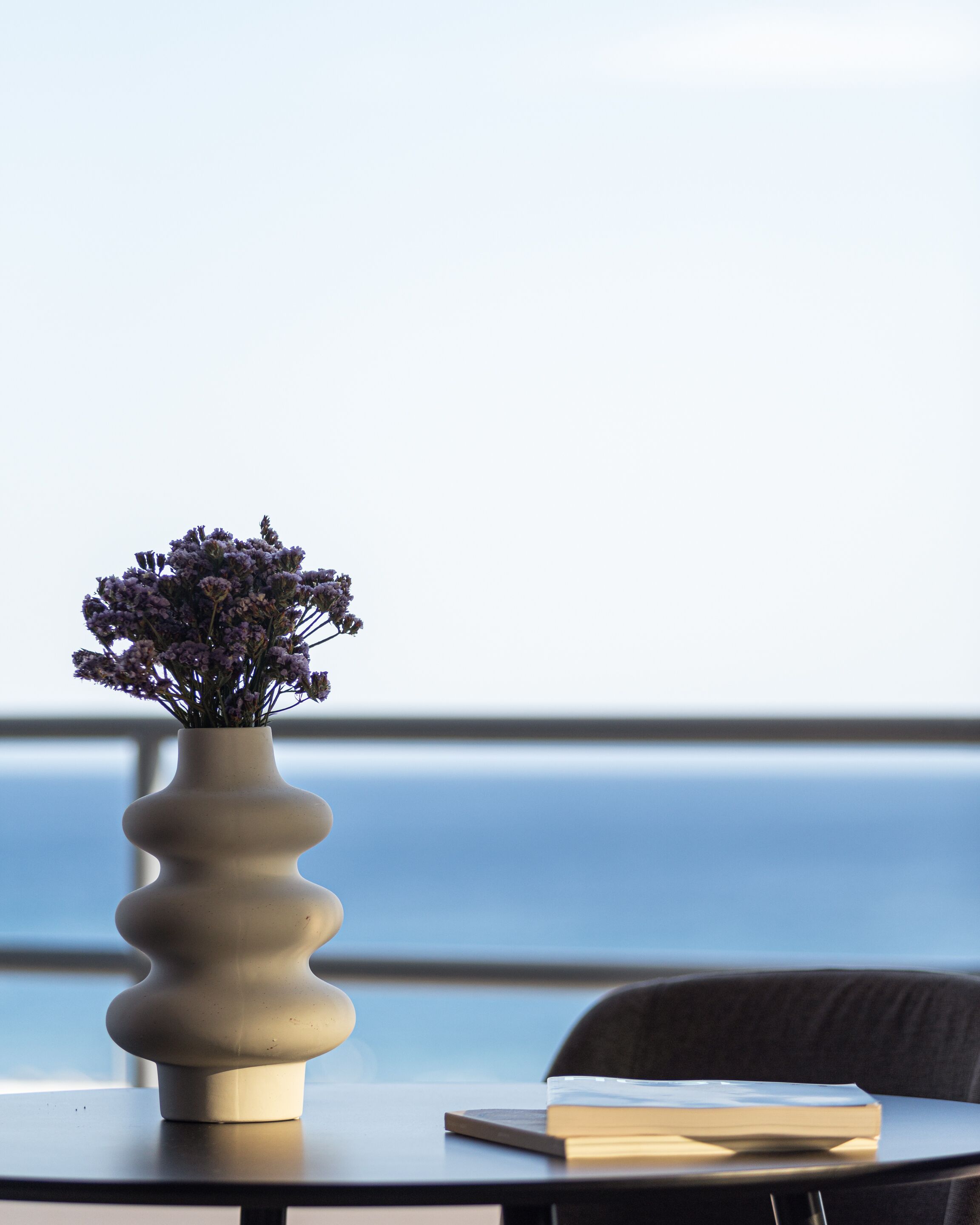 Una mesa con un florero y un libro, con vistas a un cuerpo de agua.