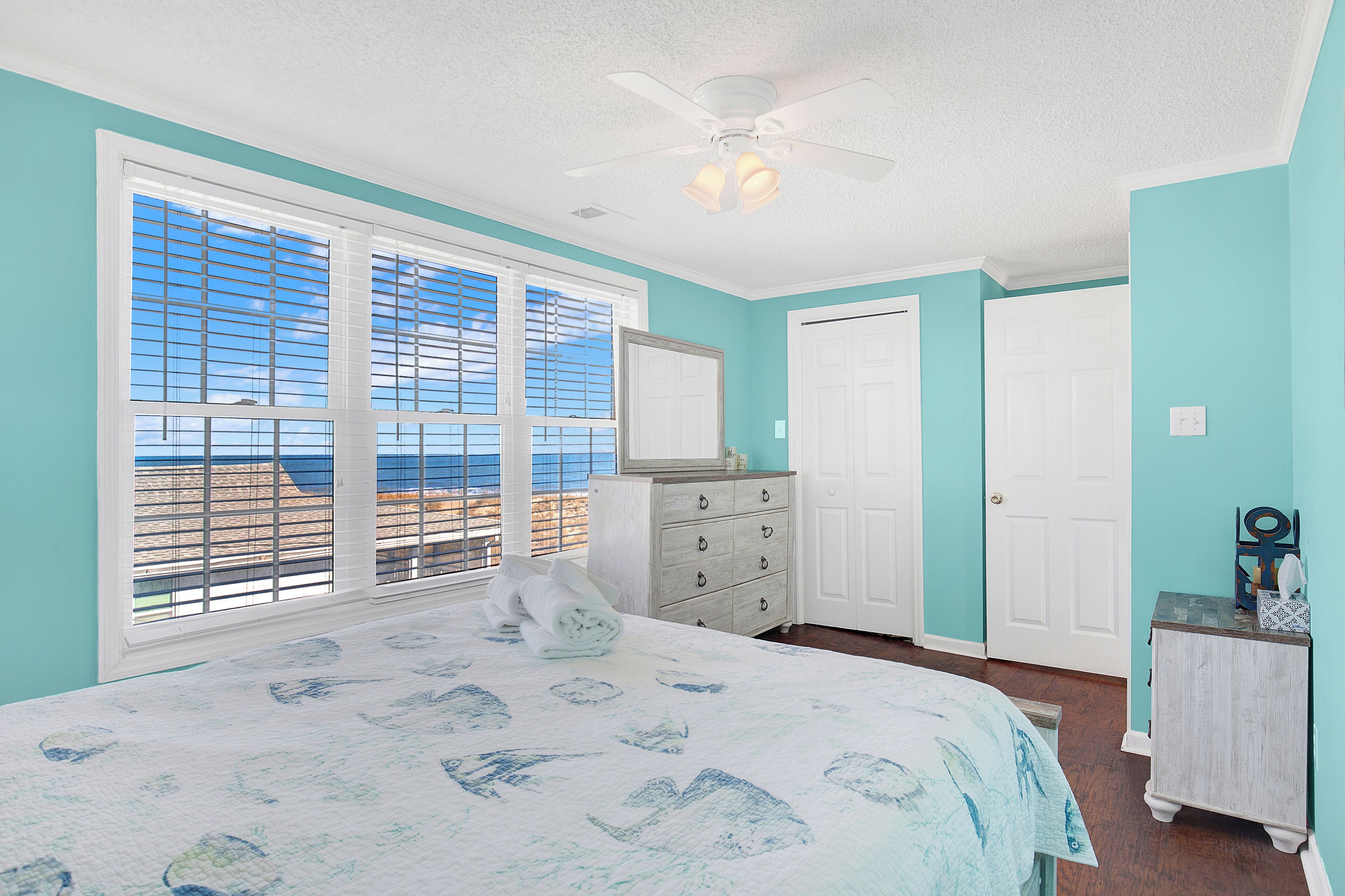 A bedroom with a large bed, a dresser, a ceiling fan, and a view of the beach through the window.