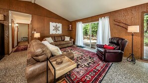 A cozy living room with a brown leather sofa, a brown armchair, and a wooden coffee table. There is a large window with white curtains, a red and black patterned rug, and a wooden wall with a snowflake decoration. - Mystic Meadow ~ Sweet Cabin + Meadow Views (Shaver Lake)