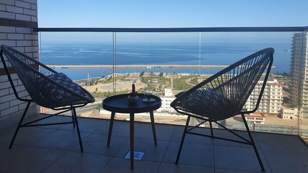 A balcony with two black metal chairs and a small round table, overlooking a coastal view with buildings and a marina. - Oran sea & bay apartment (Oran)