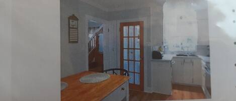 A room with a wooden table, a chair, a door, and a small kitchenette.