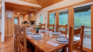 Une table de salle à manger en bois, avec des chaises, pour six personnes, une cuisine en arrière-plan et de grandes fenêtres donnant sur une zone boisée.