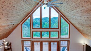 Une pièce spacieuse avec un plafond en pente, de grandes fenêtres donnant sur un paysage boisé, un ventilateur de plafond et une terrasse en bois visible à travers les fenêtres.
