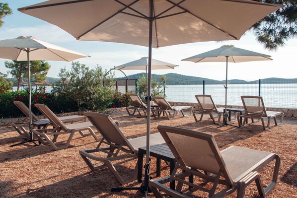 A beachside lounge area with sun loungers and umbrellas, overlooking a body of water and hills. - Beachfront Sunset Villa (Brodarica)