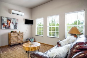 A living room with a leather sofa, a round wooden coffee table, a painting, a television, and a window with blinds.