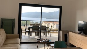 A modern living room with a sofa, a wooden dining table, and a TV. A balcony with outdoor furniture and a view of the sea and mountains is visible through large glass doors. - 10 min from the beach and 5min from the town center (Propriano)