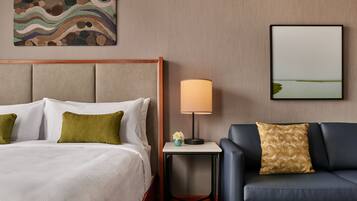 Una habitación de hotel con una cama, una mesita de noche con una lámpara, un sofá, una pared con un cuadro, y un marco con una fotografía.