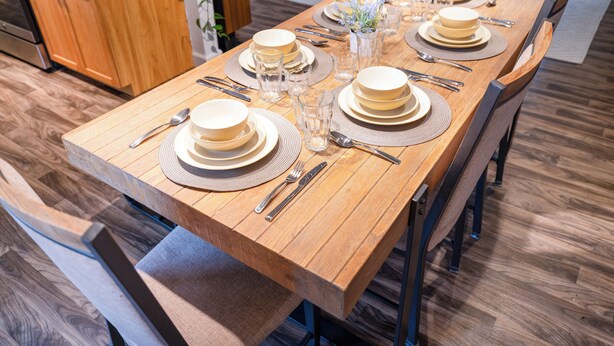 Une table à manger dressée avec des assiettes, des bols, des couverts et des verres, avec une cuisine et un coin salon en arrière-plan.