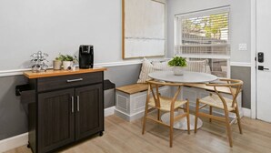 A compact kitchen with a dark wooden cabinet, a coffee maker, and a small plant on the countertop. There is a round table with chairs and a bench with cushions. A window with blinds is visible, and a door with a lock is on the right.
