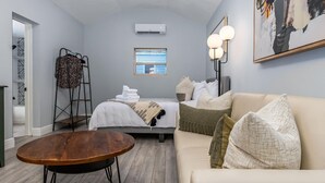 Una habitación de hotel moderna con piso de madera, un sofá color beige, una mesa de centro redonda, una cama con ropa blanca, un calefactor portátil y un cuadro abstracto de gran tamaño en la pared.