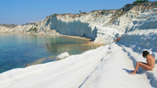 A beach with white sand and clear blue water, a person sitting on the sand, and cliffs in the background.