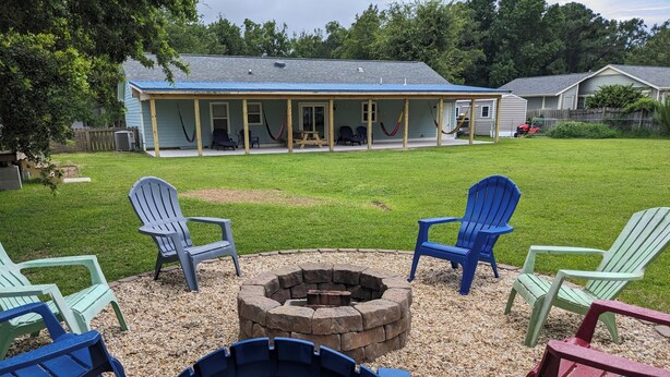 An outdoor seating area with Adirondack chairs arranged around a fire pit, a covered porch, and a lawn in the background. - Cozy Home in Central Morehead 5min. from the Beach (Morehead City)