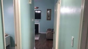 A narrow hallway with light blue walls and dark wooden flooring. There is a door on the left, a small seating area with a chair and table in the center, and a television mounted on the wall. - Cozy Home in Central Morehead 5min. from the Beach (Morehead City)