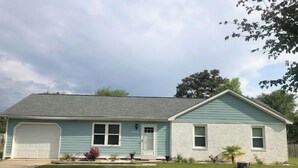 A single-story house with a garage, a front porch, and a small front yard with a lawn and a few plants. - Cozy Home in Central Morehead 5min. from the Beach (Morehead City)
