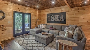 A cozy log cabin interior with a sectional sofa, a round coffee table, and a large window with a view of greenery.