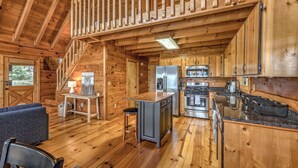 Interior de una cabaña con paredes de madera, una cocina con electrodomésticos modernos, una zona de comedor con mesa y sillas, y una escalera que sube a un nivel superior.