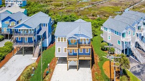 Ett bostadsområde vid kusten med flera blå hus, några med balkonger, och utsikt över havet och ett våtmarksområde i bakgrunden.