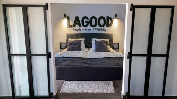 Une chambre à coucher avec un lit, des oreillers, un tapis et un panneau mural sur lequel on peut lire : « LAGOOD, les gens, la planète, la prospérité ».
