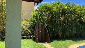 A pool area with a pathway leading to a wooden gate, surrounded by lush greenery and palm trees. - Beach House Vacation Rental 15 to 22 July 2022 Costa do Sauípe BA (Mata de São João)