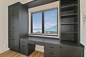 Quarto com escrivaninha embutida, janela com persianas e vista para um edifício.