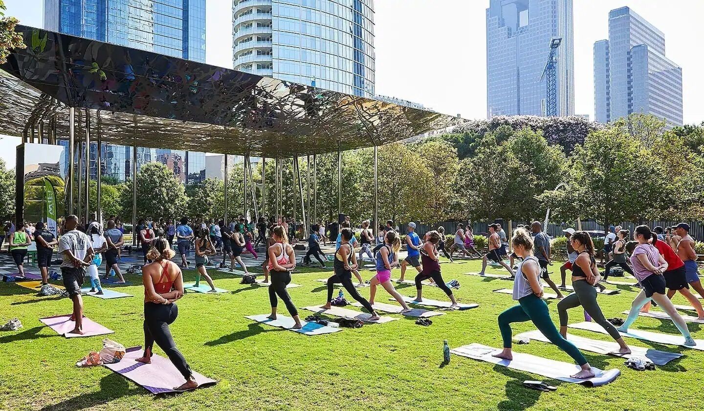 A group of people doing yoga on mats in an outdoor park with buildings in the background.