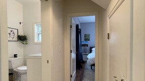 Un couloir au plancher de bois mène à une chambre à coucher où se trouvent un lit et une table de chevet. On aperçoit, sur la gauche, une salle de bain avec des toilettes et un lavabo.