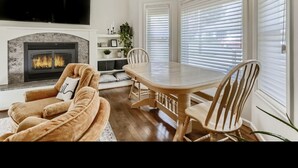 Un salon moderne doté d’une cheminée, d’un téléviseur à écran plat, d’une table à manger avec des chaises et de grandes fenêtres équipées de stores.