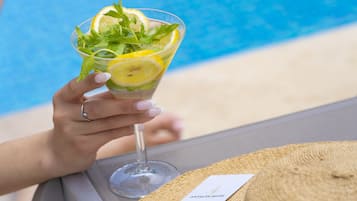 Un cocktail con fette di limone e foglie di menta, tenuto da una mano con un anello, accanto a un cappello di paglia e a un biglietto su un tavolo vicino a una piscina.