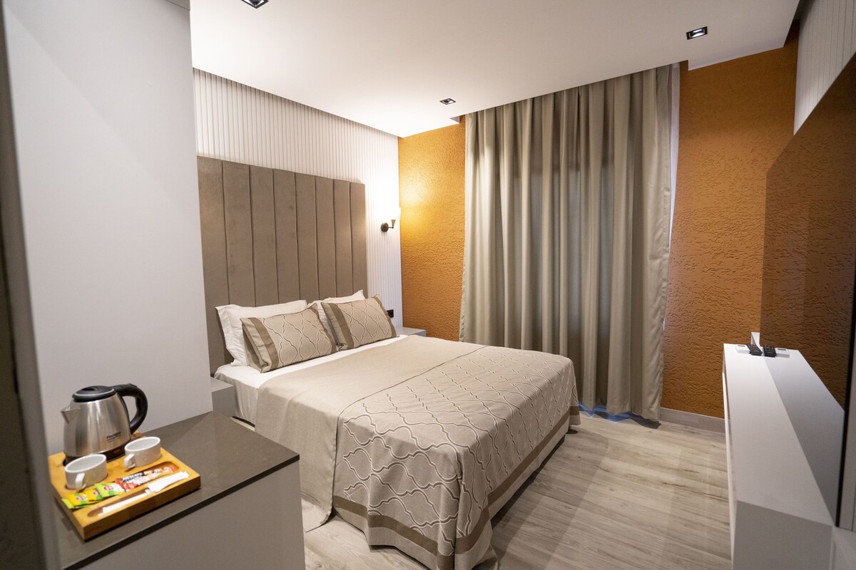 A hotel room with a bed, bedside table, and a desk with a kettle and cups.