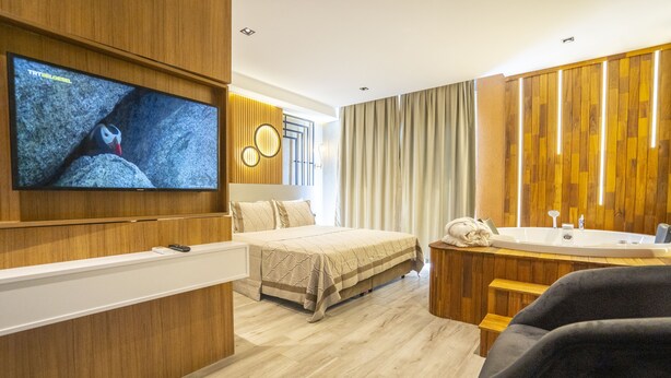Ein modernes Hotelzimmer mit einem großen Bett, einem an der Wand montierten Fernseher, einer Holztischlerei mit Waschbecken und einem großen Fenster mit Vorhängen.
