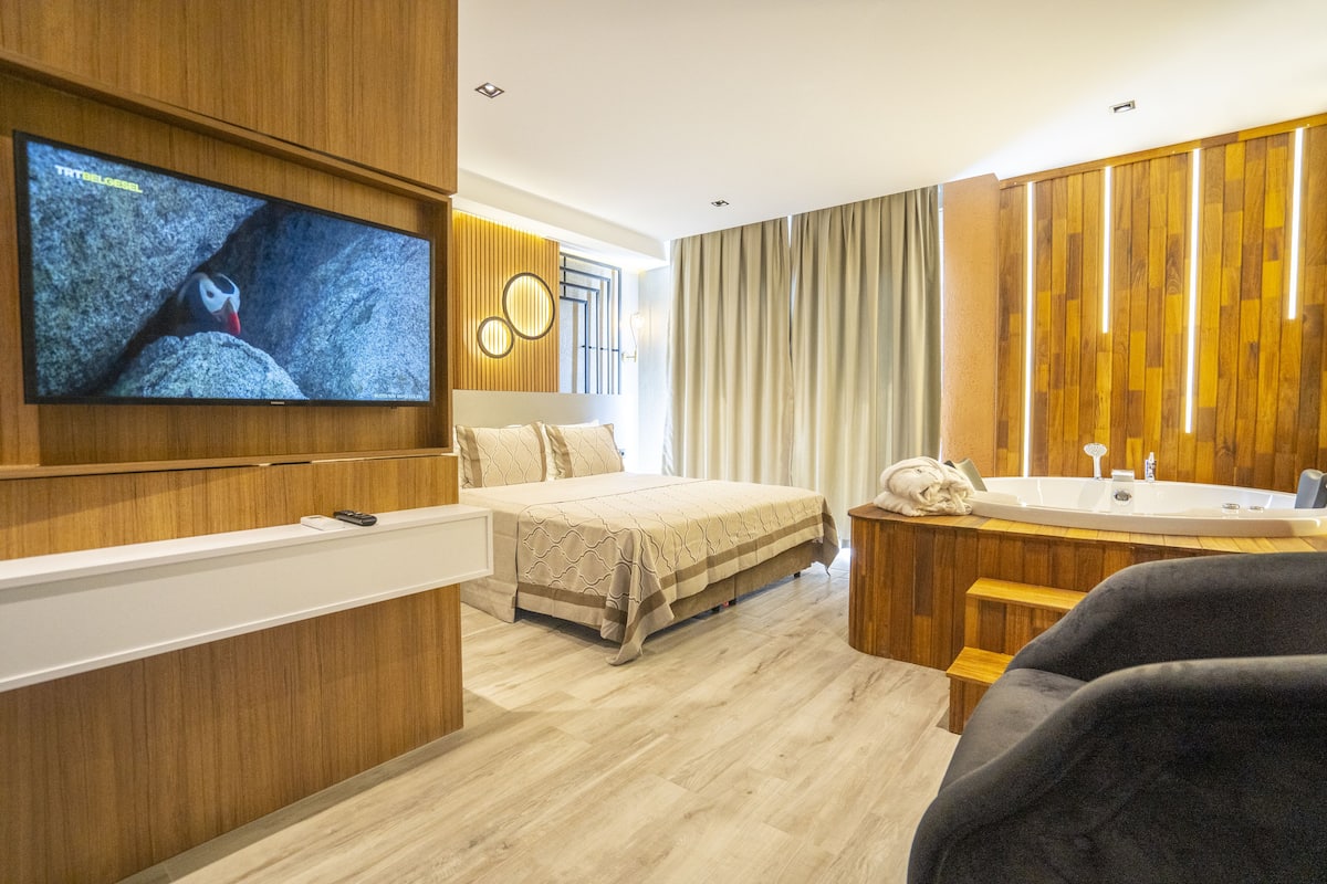 Een moderne hotelkamer met een groot bed, een aan de muur bevestigde televisie, een houten wastafel met een wasbak en een groot raam met gordijnen.