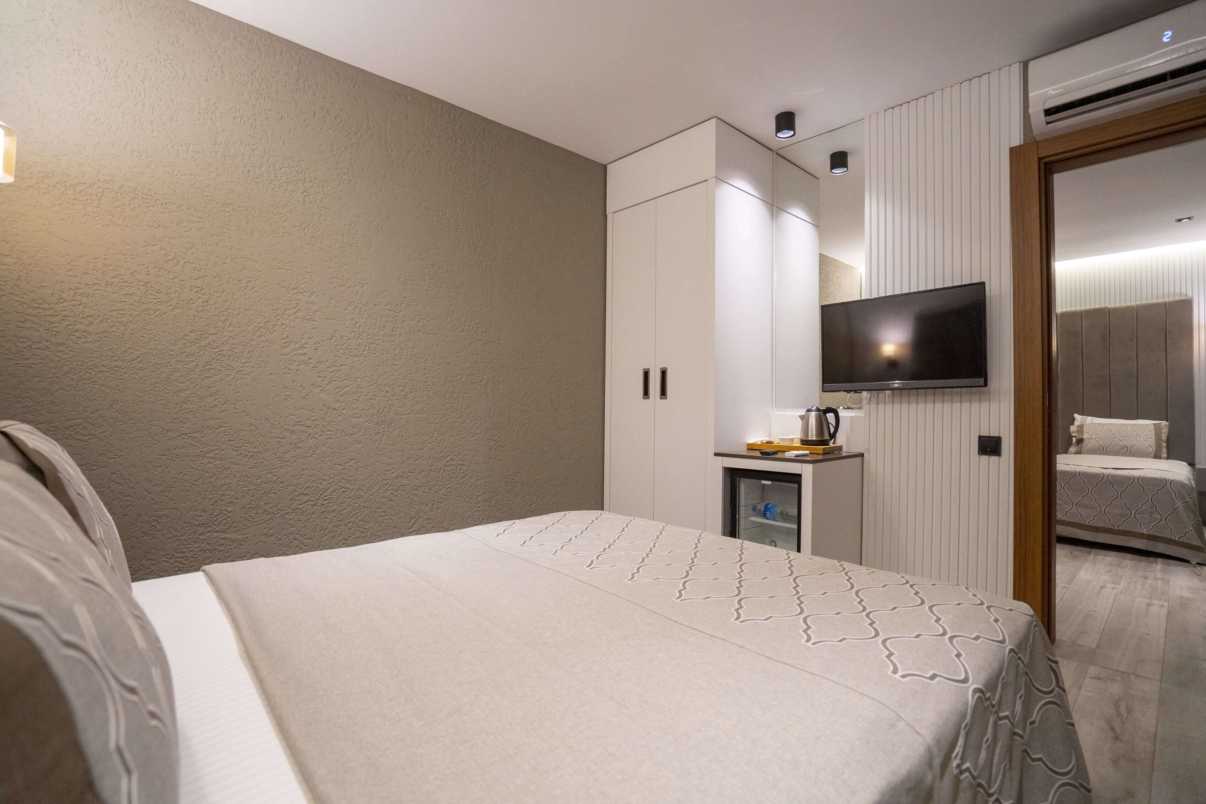A hotel room with a bed, a TV mounted on the wall, a small table with a kettle, and a wardrobe.