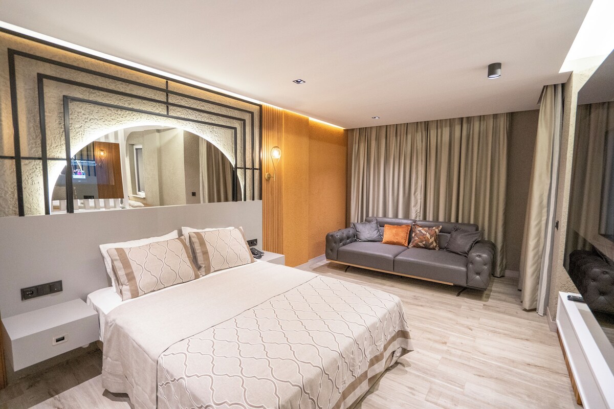 A modern hotel room with a large bed, a sofa, a TV, and a large mirror.