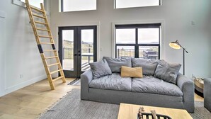 Un salon moderne avec un canapé gris, une échelle en bois et de grandes fenêtres donnant sur l’extérieur.