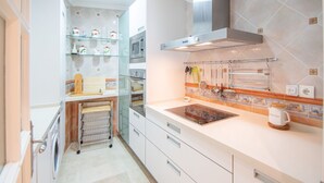 A compact kitchen with white cabinets, a microwave, a built-in oven, a glass shelf with kitchen items, and a coffee machine. - Cadiz Meridian Apartment (Cádiz)