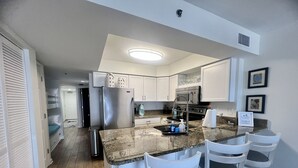 A modern kitchen with a granite countertop, stainless steel appliances, and white cabinetry. There are three white bar stools and a stainless steel sink.