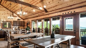Ein großzügiger Essbereich aus Holz mit mehreren Tischen und Bänken, große Fenster mit Blick ins Freie und eine Küchenzeile mit Holdekor und einer Pendelleuchte.