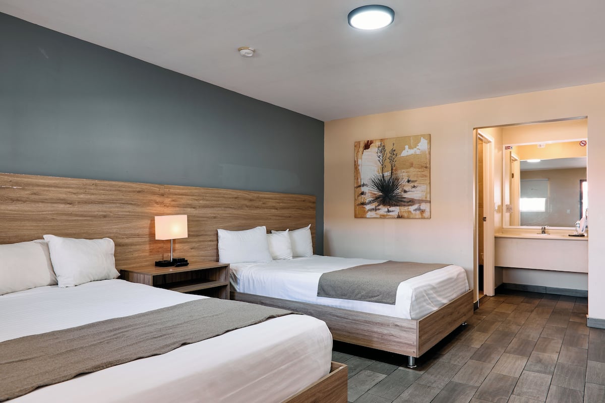 Habitación de hotel con dos camas, una mesita de noche con lámpara, un cuadro en la pared y un baño visible a través de una puerta abierta.