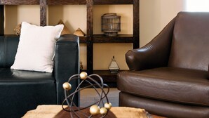 A living room with a black leather sofa, a brown leather armchair, a wooden coffee table with a decorative metal sphere, and a wooden shelving unit with various decorative items. - Hotel Dunas (Ciudad Juárez)