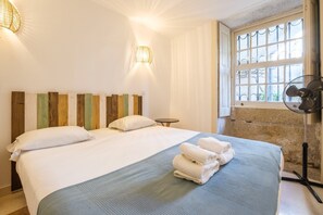 Une chambre d’hôtel avec un lit, une petite table, un ventilateur sur pied et une fenêtre donnant sur un beau paysage.