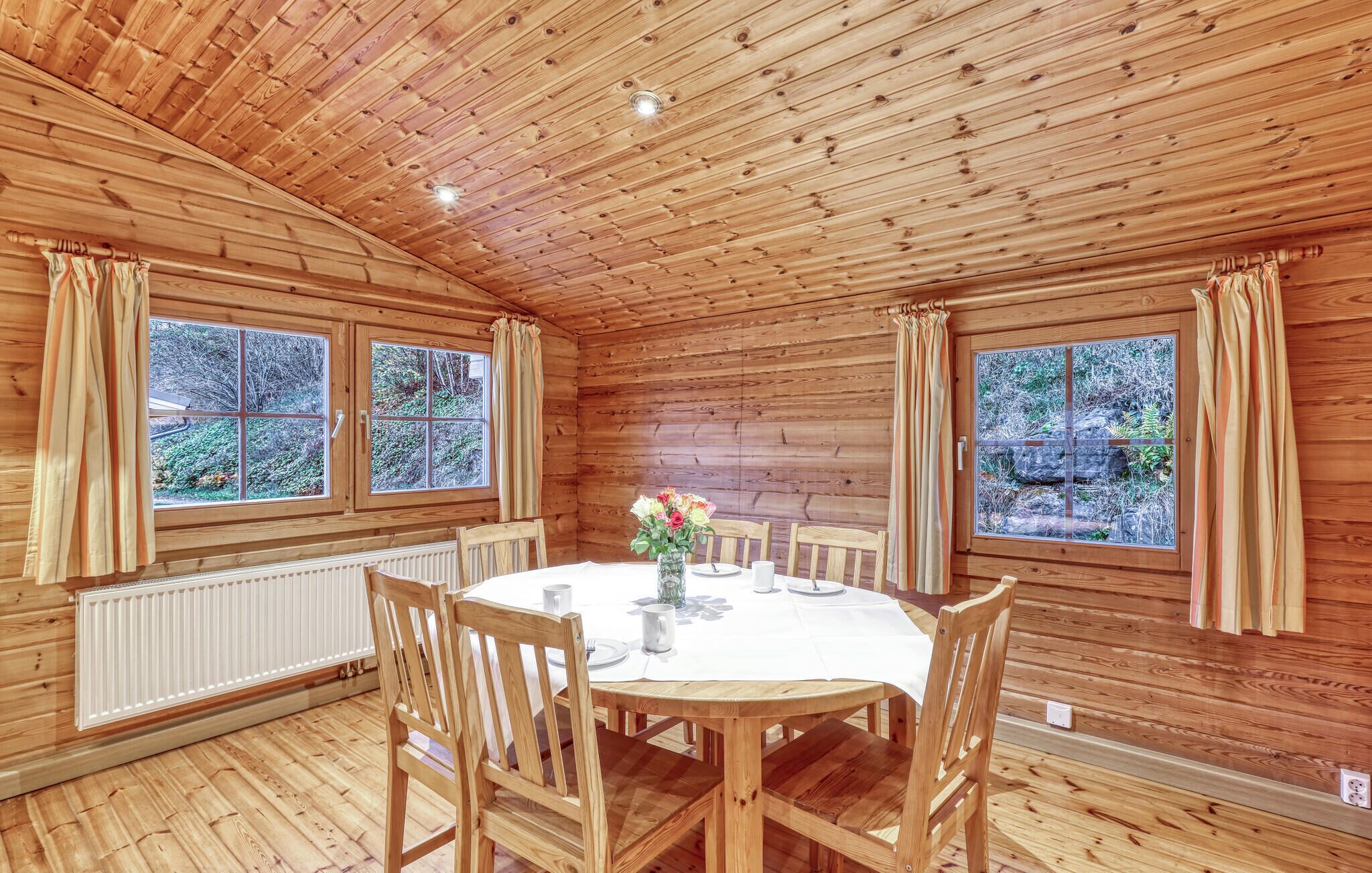 A wooden cabin with a round dining table, four chairs, a vase with flowers, and two windows with curtains.