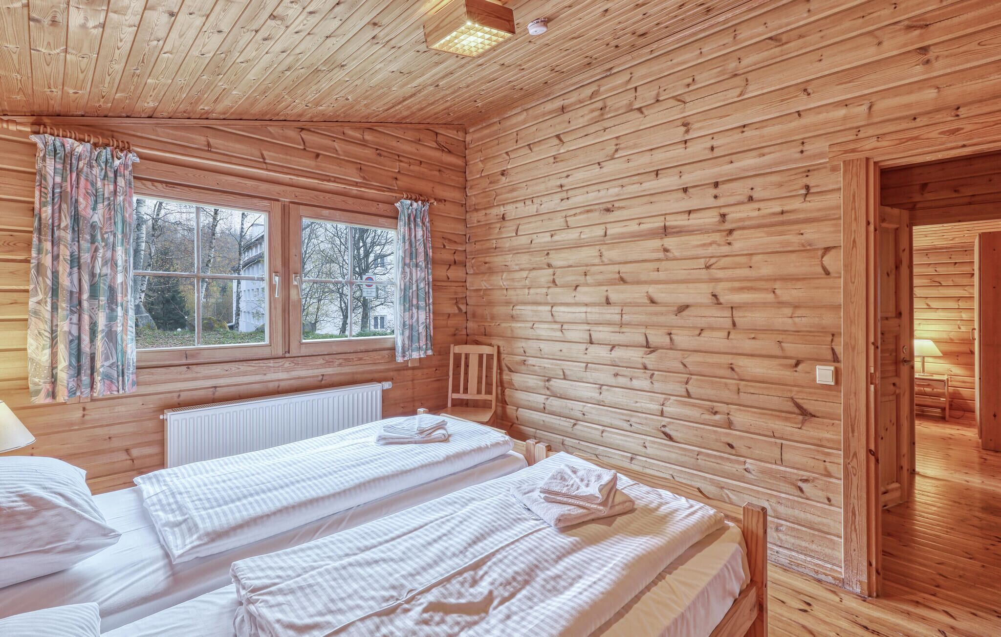 Een slaapkamer in een houten chalet met twee bedden, een raam met gordijnen en een radiator.