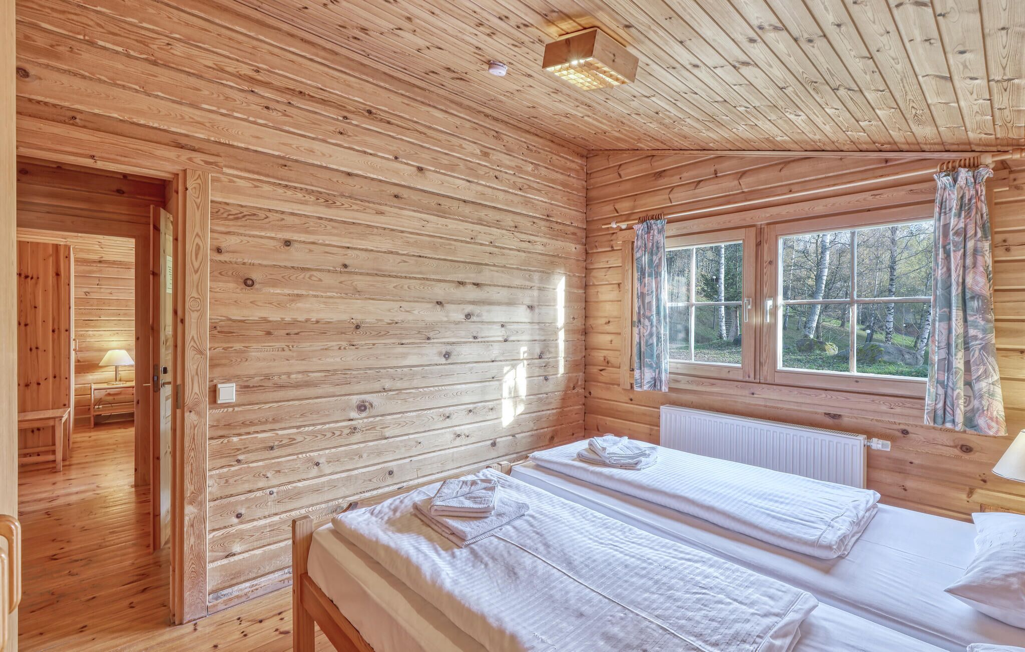 En stuga med träpanel, två sängar, ett fönster med gardiner och en dörr som leder till ett annat rum.