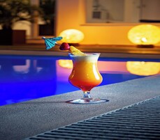 Un cóctel en un vaso con pajita y una cereza sobre una sombrilla, colocado junto a una piscina con otra piscina al fondo.