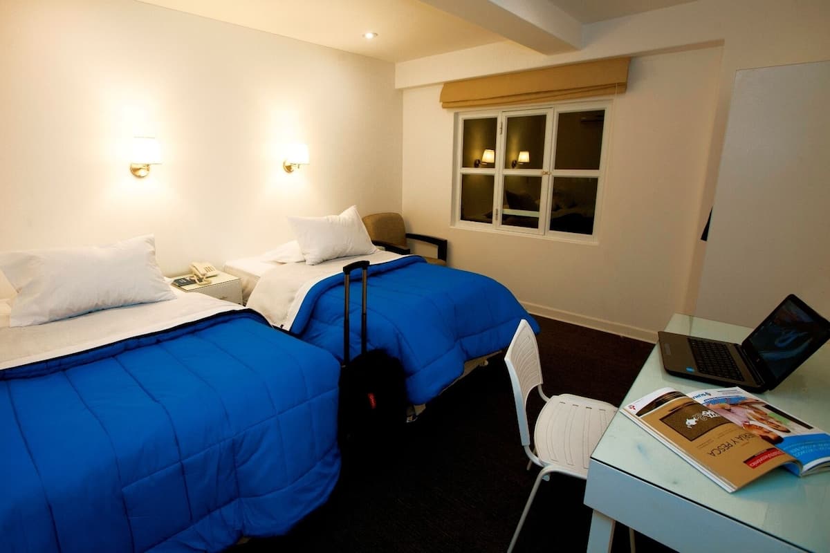 Quarto de hotel com duas camas, uma escrivaninha com um laptop e uma janela com cortinas.