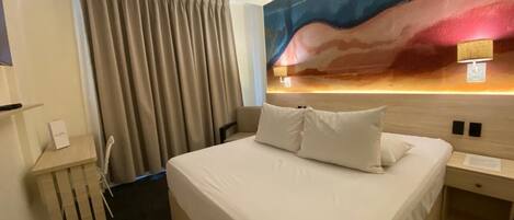 Habitación de hotel con una cama grande, cabecero con pintura, mesita de noche y una ventana con cortinas.