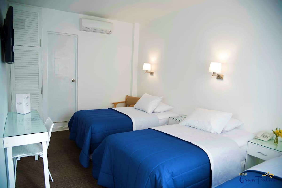 Quarto de hotel com duas camas, uma escrivaninha, uma cadeira, mesa de cabeceira com abajur e televisão.