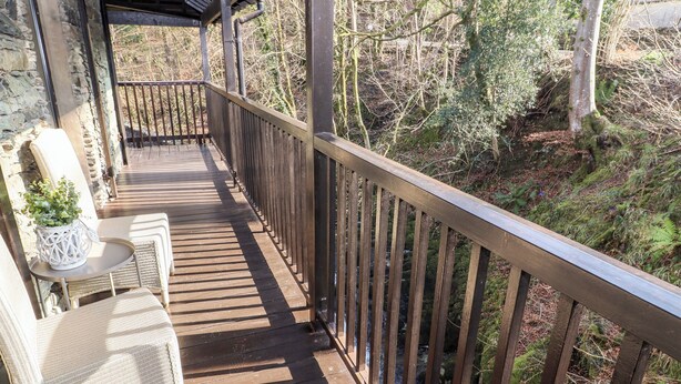 A wooden deck with wicker chairs and a table, overlooking a forested area and a stream. - Leafy Nook (Ambleside)
