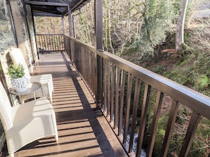 A wooden deck with wicker chairs and a table, overlooking a forested area and a stream. - Leafy Nook (Ambleside)